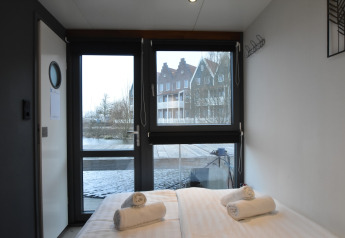 Bedroom on houseboat 'Boat' in the Netherlands with a water and houses view through large windows.