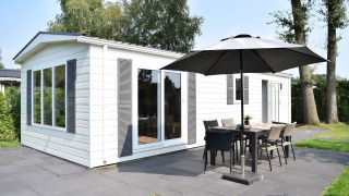 Veluwelodge bij Vakantiepark De Boshoek in Nederland met witte gevel, terras, eettafel en grote parasol buiten.