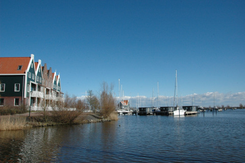 Panorama of a canal with a houseboat named 'Boat', yachts, and Dutch houses under a clear blue sky.