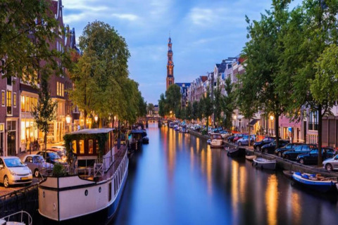 Photo of a houseboat named 'Boat' on a Dutch canal, surrounded by cars, trees, and lit buildings at dusk.