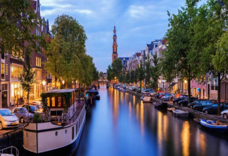 Photo of a houseboat named 'Boat' on a Dutch canal, surrounded by cars, trees, and lit buildings at dusk.