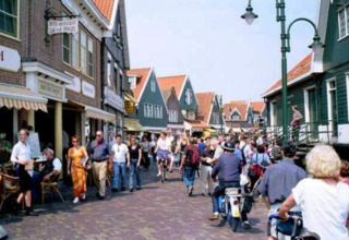 Busy Dutch street scene with people walking, cycling, and riding scooters past traditional houses.