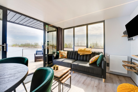 Soggiorno sulla houseboat 'La Belle Vie' in Olanda, arredamento moderno, grandi finestre e vista sull’acqua.