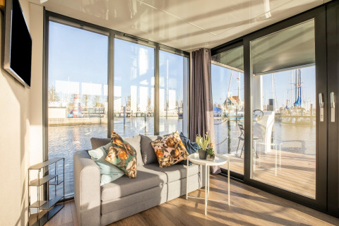 Stue med sofa og udsigt over vandet på husbåden Marina Sunrise i Holland, med store vinduer og terrasse.