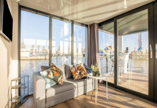 Stue med sofa og udsigt over vandet på husbåden Marina Sunrise i Holland, med store vinduer og terrasse.