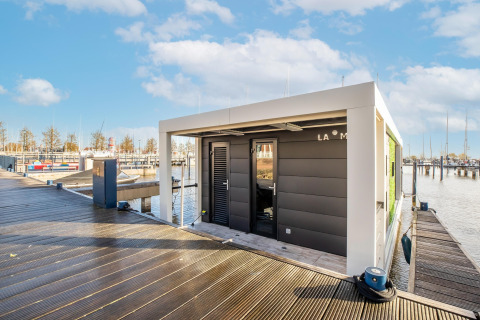 Moderne husbåd ved en marina i Nederlandene på en solskinsdag, set fra en træmole med både i baggrunden.
