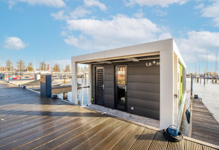 Moderne husbåd ved en marina i Nederlandene på en solskinsdag, set fra en træmole med både i baggrunden.