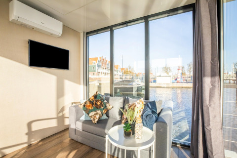 Lyst stueområde på husbåden ‘Marina Sunrise’ i Holland, med sofa, dekorationer og udsigt til vandet.