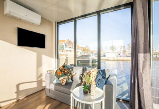 Lyst stueområde på husbåden ‘Marina Sunrise’ i Holland, med sofa, dekorationer og udsigt til vandet.