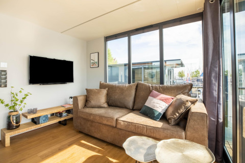 Stue med stor brun sofa, fladskærms-tv, og store vinduer på husbåd i Volendam Marina, Holland.