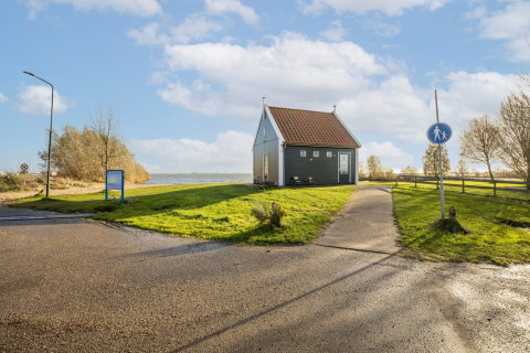 Et lille husbåd ved vandet, omgivet af grønt græs og træer, med blå himmel og skyer i Holland.