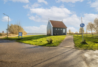 Et lille husbåd ved vandet, omgivet af grønt græs og træer, med blå himmel og skyer i Holland.