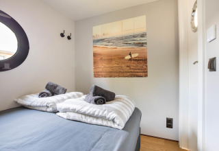 Soveværelse i husbåd i Volendam Marina, Holland, med dobbeltseng, surfebillede og runde vinduer.