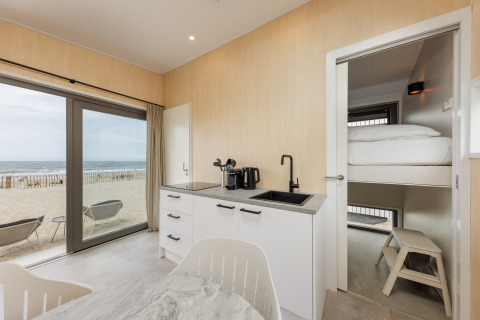 Moderne strandhusinteriør med køkken, køjeseng og havudsigt gennem store vinduer ved stranden.