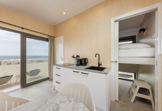 Moderne strandhusinteriør med køkken, køjeseng og havudsigt gennem store vinduer ved stranden.