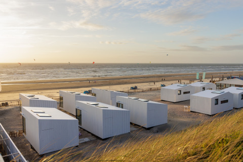 Moderne strandhytter med hvid facade og store vinduer står på sandstrand ved havet ved solnedgang.