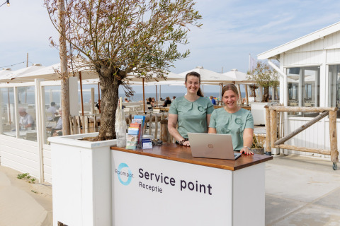 To smilende kvinder står ved en servicepoint-reception foran et strandhus på en solskinsdag.