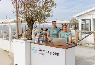 To smilende kvinder står ved en servicepoint-reception foran et strandhus på en solskinsdag.