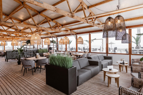 Lyst og moderne strandhusinteriør med træloft, rattanlamper og komfortable siddeområder ved havet.