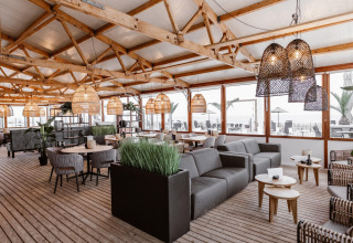 Lyst og moderne strandhusinteriør med træloft, rattanlamper og komfortable siddeområder ved havet.
