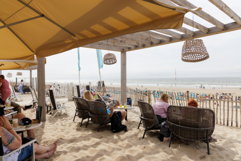 Folk slapper af under en overdækning på en strandcafé, med udsigt til havet og folk på stranden.