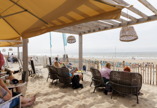 Folk slapper af under en overdækning på en strandcafé, med udsigt til havet og folk på stranden.