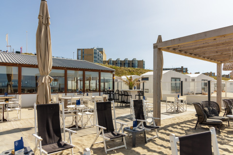 Udeservering ved strandhus med stole, borde, sand og udsigt til moderne bygninger i baggrunden.