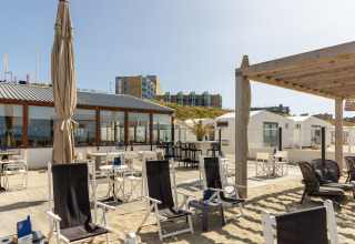 Udeservering ved strandhus med stole, borde, sand og udsigt til moderne bygninger i baggrunden.