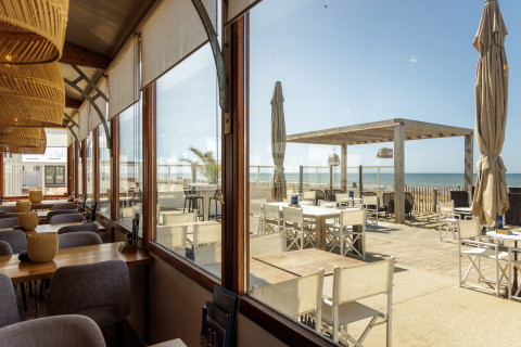 Indendørs og udendørs siddepladser ved en strandcafé med udsigt over havet gennem store vinduer.