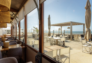 Indendørs og udendørs siddepladser ved en strandcafé med udsigt over havet gennem store vinduer.