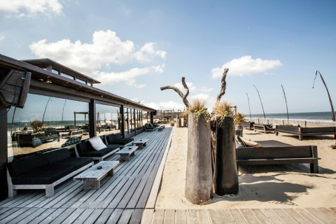 Udsigt over en moderne strandcafé med træterrasse og loungeområder, direkte ved havet på en solrig dag.