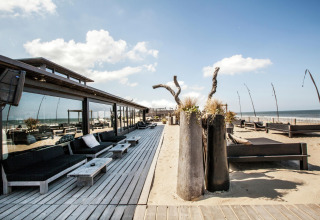 Udsigt over en moderne strandcafé med træterrasse og loungeområder, direkte ved havet på en solrig dag.