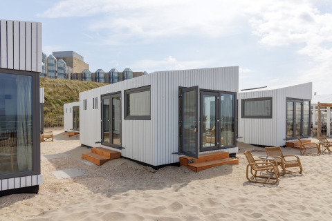 Strandhuse Zandvoort 3 i Holland med moderne hvide hytter, træstole og udsigt til klitter og bygninger.