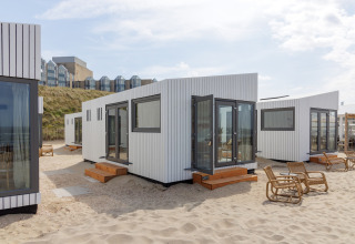Strandhuse Zandvoort 3 i Holland med moderne hvide hytter, træstole og udsigt til klitter og bygninger.