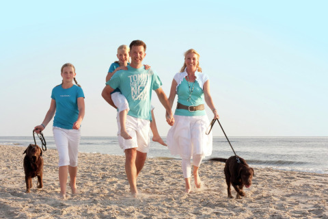 En familie med to børn og to hunde går langs stranden ved et sommerhus på en solrig dag.