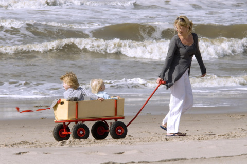 Kvinde trækker en vogn med to små børn langs sandstranden ved havet med bølger i baggrunden.