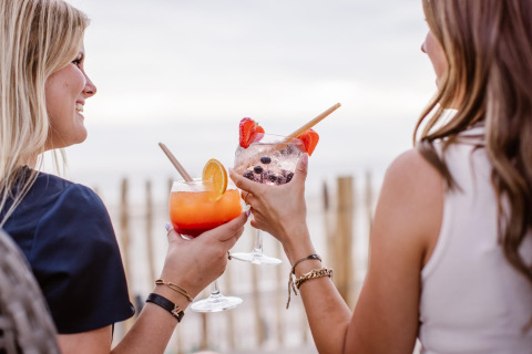 To kvinder nyder farverige cocktails ved stranden med udsigt over havet, smilende og afslappede.