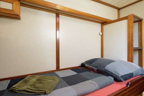 Eenpersoonsbed op boot in Leiden met houten details, geruit beddengoed en handdoeken op het bed.