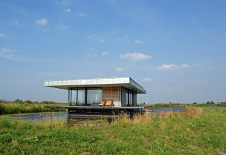Moderne husbåd, Watervilla de Blauwe Reiger, på stille vandvej i Holland med blå himmel og græs.