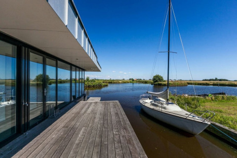 Moderne husbåd ved vandet med stor træterrasse og sejlbåd i smukke omgivelser i Nederlandene.