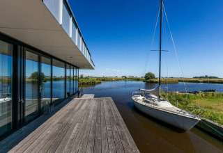 Moderne husbåd ved vandet med stor træterrasse og sejlbåd i smukke omgivelser i Nederlandene.