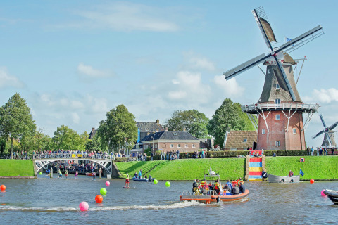 Festlig scene ved en kanal med husbåd, vindmølle, farverige bolde og folk i både i Holland.