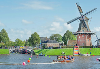Festlig scene ved en kanal med husbåd, vindmølle, farverige bolde og folk i både i Holland.