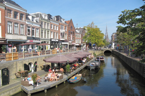 Hus både ved kanalen med folk der nyder caféer og solskin, Watervilla de Blauwe Reiger, Holland.