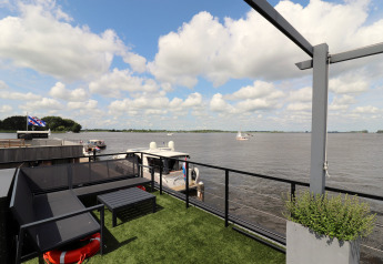 Udsigt fra husbåd Kohlenpott i Holland, med terrasse, sofa og udsigt over floden med både og skyer.