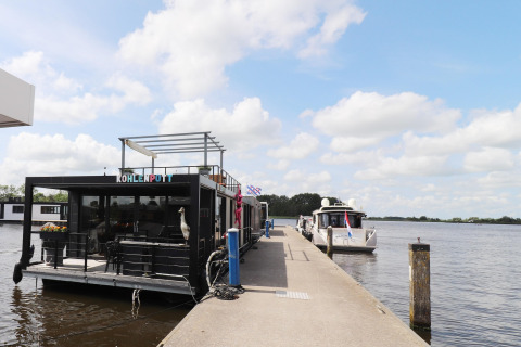 Casa flotante 'Kohlenpott' atracada en un muelle en Países Bajos, con barcos y cielo parcialmente nublado.