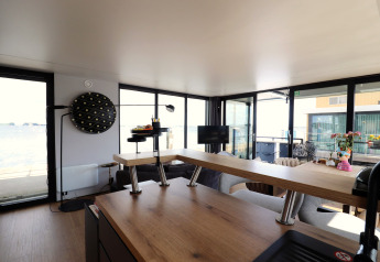 Stue på husbåd med moderne interiør, store vinduer og udsigt over vandet og terrasse i Holland.