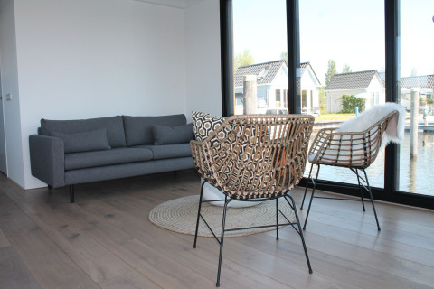 Moderne woonkamer op een woonboot in Stavoren, Nederland, met grote ramen, grijze bank en rotan stoelen.