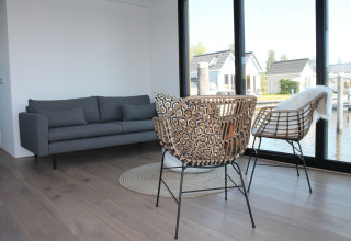Moderne woonkamer op een woonboot in Stavoren, Nederland, met grote ramen, grijze bank en rotan stoelen.
