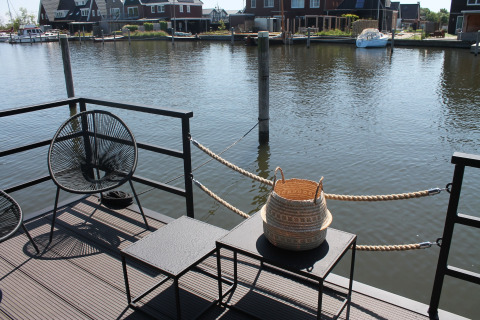 Uitzicht vanaf een woonboot bij Boat House Stavoren, Nederland, met stoelen, tafels en een mand aan het water.